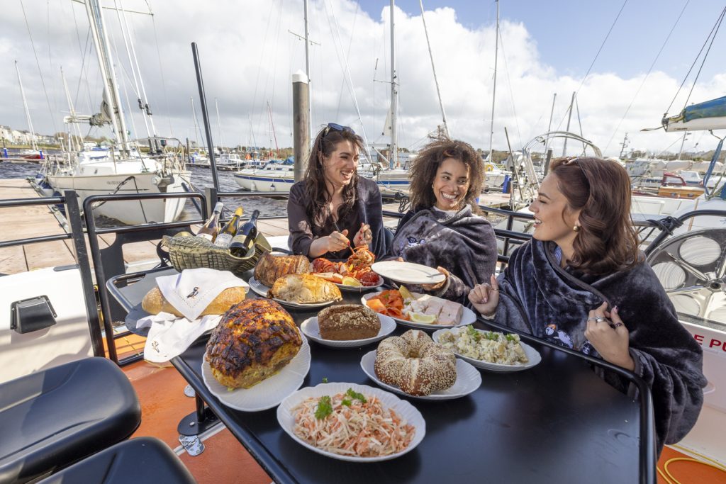 Guests seated around a table on deck laden with seafood platters, breads and salads, moored among yachts