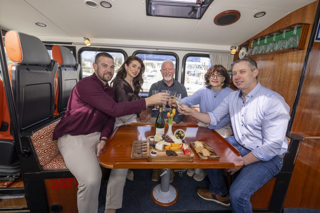 Five guests around the cabin table of Puifín raising their glasses, with a cheese and charcuterie board and Champagne in front of them