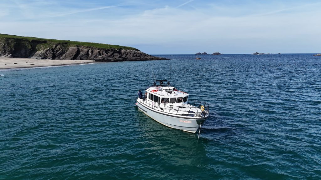 Puifín at anchor in clear blue water beside a sheltered sandy cove under low cliffs
