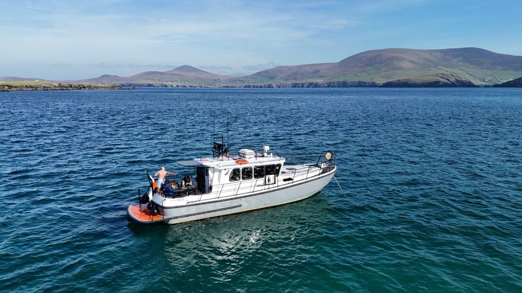 Guests standing on the rear deck of Puifín in calm blue water, with the mountains of the Dingle Peninsula behind
